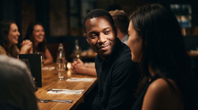 A smiling man at a lively dinner table looking tenderly at the woman beside him