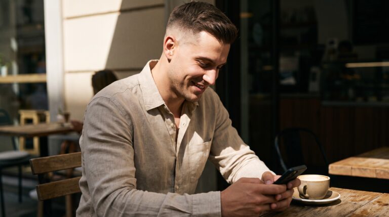 A stylish man smiling at his phone while texting on a sunny cafe terrace